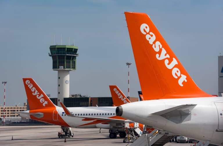 A lineup of EasyJet airplanes parked on the tarmac at Newquay airport, each with distinctive orange tails featuring the EasyJet logo. In the background, an air traffic control tower rises behind the aircraft, set against a hazy sky. The scene captures a typical busy airport environment with a focus on commercial aviation and travel infrastructure.