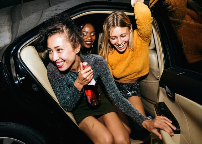 Late night taxi Newquay.Two stylish individuals seated in the back of a beige car, surrounded by a sleek interior. One person, dressed in a sparkly long-sleeved top and short skirt, holds a bottle while facing slightly toward the camera. The other, wearing a yellow sweater and sequined skirt, leans toward the car door, appearing to exit or adjust something. The mood evokes a sense of nightlife energy or post-party ambiance, with fashion-forward flair and intimate framing. Let me know if you'd like a version that feels more editorial, playful, or tailored for specific platforms like Instagram or accessibility tools.