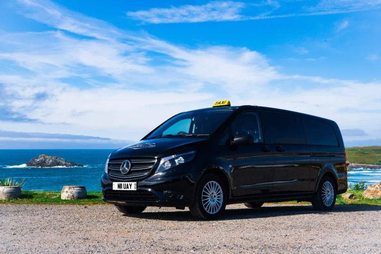 Group travel in Newquay. Black Mercedes-Benz taxi van parked on a gravel surface near a scenic coastline in Newquay. A “TAXI” sign sits atop the van, and the license plate reads “N11 UAY.” In the background, the ocean stretches to the horizon with a small rocky island and a partly cloudy sky.
