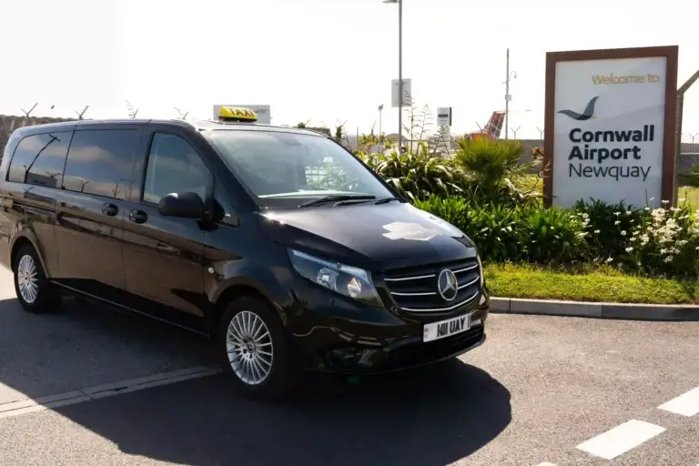 Newquay Airport Taxis. Black Mercedes-Benz taxi van parked beside a sign that reads “Welcome to Cornwall Airport Newquay.” The van features a “TAXI” sign on its roof. In the background, airport facilities and greenery suggest the entrance area to the airport.