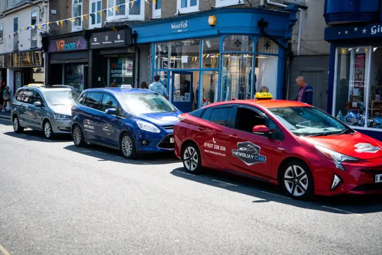 Street view featuring three Newquay Cab taxis parked in a row—red, blue, and silver—each with roof signs and matching branding. The red taxi prominently displays the phone number “01637 226 224” and the website “www.newquaycab.com .” Behind the vehicles, shops including “VRcade” and “Weird Fish” are visible, with pedestrians strolling along the sidewalk, adding a touch of local charm to the busy seaside town atmosphere.
