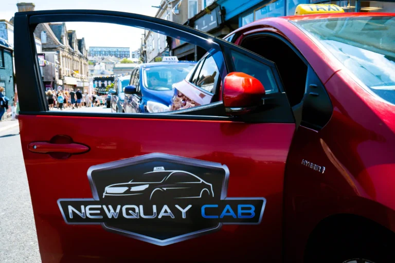 Red hybrid taxi branded “NEWQUAY CAB” parked along a bustling street lined with shops and pedestrians. The cab features a yellow roof sign, logo on the door, and a “HYBRID” badge, suggesting eco-conscious design. In the lively background, more vehicles and people add motion and energy to this vibrant commercial area.